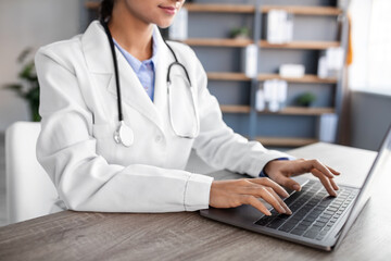 Wall Mural - Cropped young hindu woman doctor in white coat with stethoscope typing on computer in office interior