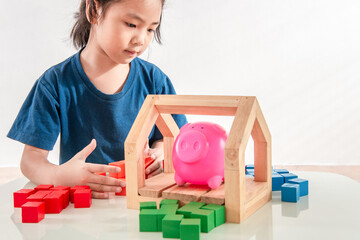 A happy little girl playing with colorful toy blocks wooden house with a pink piggy bank to creates constructions. Money saved in the future, learning game for childhood baby and toddler idea