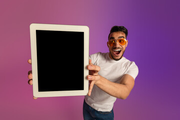 Poster - Excited millennial Arab man in sunglasses showing tablet with blank screen in neon light, mockup for app or website