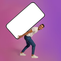 Wall Mural - Full length of millennial Arab guy carrying big heavy smartphone with empty white screen in neon light. Mockup