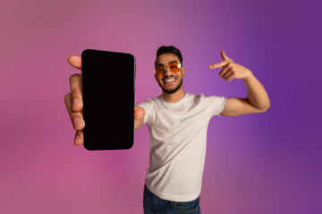 Poster - Cool young Arab guy pointing at cellphone with empty black screen, recommending new app or website in neon light, mockup