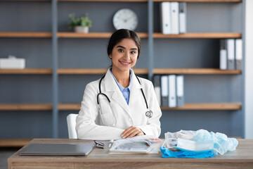 Smiling pretty young hindu woman doctor in white coat with stethoscope sits at table with many protective mask