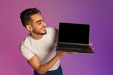 Poster - Millennial Arab man showing laptop computer with empty black screen in neon light, mockup for new website