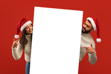 Place For Ad. Happy Middle Eastern Couple In Santa Hats Holding White Board, Looking And Pointing On Empty Placard