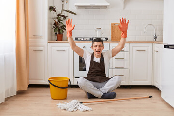 Wall Mural - Excited happy brunette Caucasian man wearing casual clothing, apron and orange rubber gloves, sitting on floor in kitchen with crossed legs, raised arms, being glad to finish cleaning house.