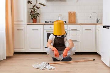 Wall Mural - Portrait of unknown anonymous male housekeeper sitting on floor in kitchen with yellow bucket on his head, cleaning house, washing floor, being tired.