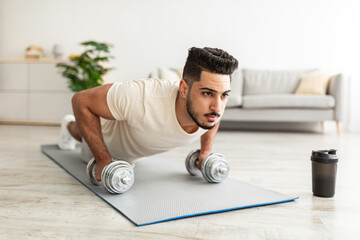 Wall Mural - Stay home sports. Handsome young Arab man standing in plank pose with dumbbells, working out indoors