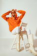 Fashionable confident woman wearing trendy orange hoodie, sunglasses, white skinny jeans, high boots, sitting, posing on chair. Full body portrait