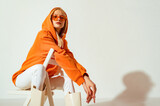 Fototapeta  - Young confident blonde girl wearing trendy orange hoodie, color sunglasses, posing on white background. Studio fashion portrait. Copy, empty space for text