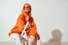 Fashionable Girl Wearing Orange Hoodie, Glasses Looking Up,  Posing On White Background. Copy, Empty Space For Text
