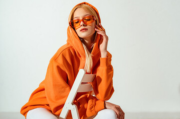 indoor fashion portrait of young confident blonde woman wearing trendy orange hoodie, color sunglass