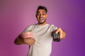 Poster - Cheerful millennial Arab guy watching TV and eating popcorn, switching channels with remote control in neon light