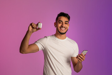 Wall Mural - Cheerful young Arab guy using smartphone, showing wireless earphones, advertising new gadget in neon light