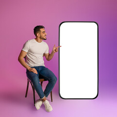 Poster - Happy young Arab guy sitting on chair, pointing at giant cellphone with empty white screen in neon light, mockup