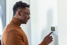 Smiling African American Man Using Modern Smart Home System, Controller On Wall, Positive Young Man Switching Temperature On Thermostat Or Activating Security Alarm In Apartment