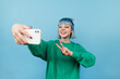 © bodnarphoto - Happy hipster girl with colored hair takes a selfie on a blue background and shows the camera a gesture of peace with a smile on his face.