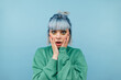 © bodnarphoto - Portrait of shocked teen girl with colored hair on blue background, looking at camera and holding cheeks, wearing green sweatshirt