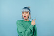 © bodnarphoto - Surprised girl with blue hair looks at the camera with a shocked face and points his finger to the side, isolated on a blue background.
