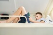 © timltv - Teen boy with short blond hair reads electronic book via tablet PC lying on white bedding on train car upper shelf close low angle shot