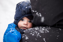 Adorable Baby In Warm Clothes In Hands Of Anonymous Parent During Winter Stroll In Snowy Weather