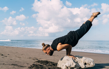 Caucasian man in casual wear doing balance pose at coastline stone spending daytime for training body strength, young male yogi with muscular hands concentrated on sportive efforts at seashore