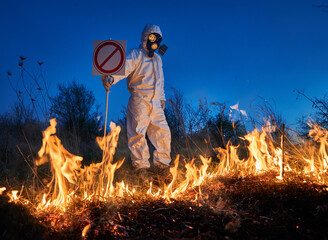 Fireman ecologist extinguishing fire in field at night. Man in protective radiation suit and gas mask near burning grass with smoke, holding prohibition sign. Natural disaster concept.