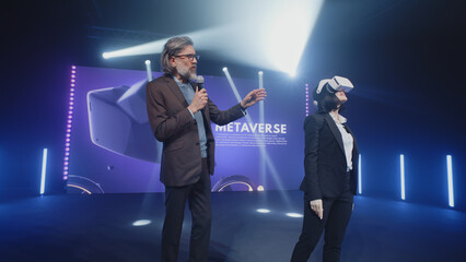 Two speakers in virtual reality glasses standing on the stage and talking about modern technologies and the development of the Metaverse, during presentation in illuminated room with an LED screen