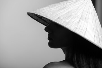 Fashion concept. Black and white portrait of beautiful woman with naked shoulder and long dark hairs wearing traditional cone-shaped Asian cane hat and cover her face in shade. Face is in focus