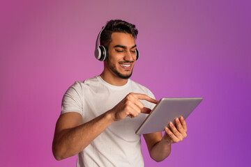 Poster - Cheery young Arab man having video call, using tablet computer, wearing wireless headphones in neon light