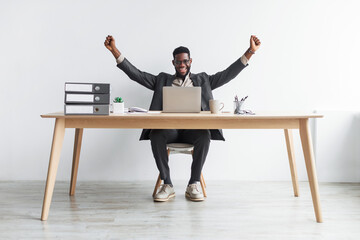 Full length of excited young black businessman sitting at desk with laptop, making YES gesture against white studio wall