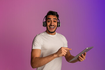 Poster - Modern gadgets for entertainment. Excited young Arab man in wireless headphones pointing at digital tablet in neon light