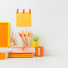 Creative desk in kids room with yellow frame mock up, plant and notebooks, note on the wall and many yellow and orange supplies.