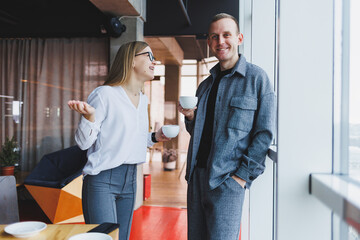 A businessman and a business woman are drinking coffee together, standing in front of the windows of an office building overlooking the city. Two confident businessmen working in a modern office