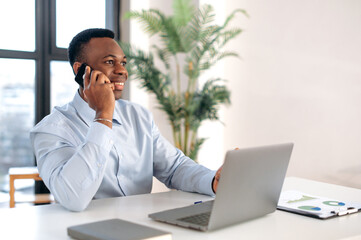 Successful confident busy African American business man, real estate agent, manager, sitting in a modern office, talking on the phone with colleagues or clients, have important conversation, smiling