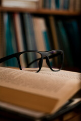 Eye glasses on top of an open book. Literature day and knowledge concept. Blurred bookshelf on the background. Selective focus.