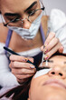 © Dusko - Beautiful young redhead woman at eyelash extension procedure. Cosmetics and body care close up shot. Female beautician wearing protective face mask against. Selective focus.