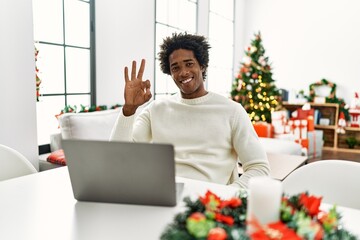 Poster - Young african american man using laptop sitting on the table by christmas tree smiling positive doing ok sign with hand and fingers. successful expression.