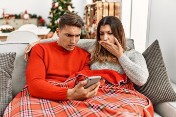 Sticker - Young hispanic couple sitting on the sofa on christmas covering mouth with hand, shocked and afraid for mistake. surprised expression