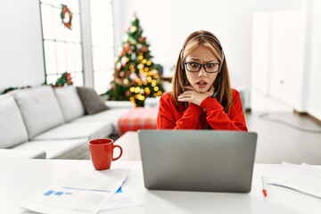 Poster - Young caucasian girl sitting on the table working using laptop by christmas tree shouting and suffocate because painful strangle. health problem. asphyxiate and suicide concept.