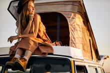 Attractive charming lady in dress and hat is on roof of yellow mini van, enjoy the sunset from top, relax at summer vacations, evening. magnificent landscape around, female in contemplation