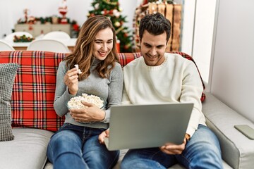 Wall Mural - Young hispanic couple watching film eating popcorn sitting on the sofa at home.