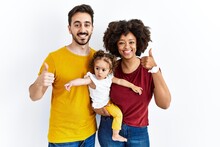Interracial Young Family Of Black Mother And Hispanic Father With Daughter Doing Happy Thumbs Up Gesture With Hand. Approving Expression Looking At The Camera Showing Success.