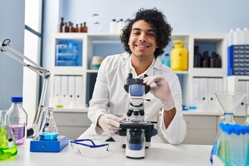 Sticker - Young hispanic man wearing scientist uniform using microscope at laboratory