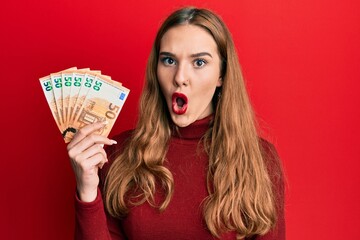 Sticker - Young blonde woman holding euro banknotes scared and amazed with open mouth for surprise, disbelief face