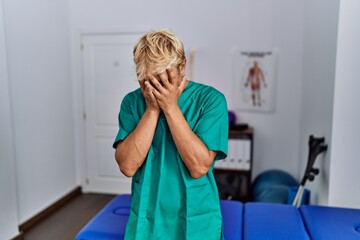 Sticker - Young blond man wearing physiotherapist uniform standing at clinic with sad expression covering face with hands while crying. depression concept.