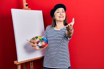 Wall Mural - Middle age hispanic woman standing drawing with palette by painter easel stand smiling friendly offering handshake as greeting and welcoming. successful business.