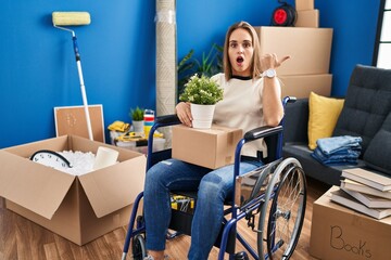 Poster - Young woman sitting on wheelchair moving to a new home surprised pointing with hand finger to the side, open mouth amazed expression.