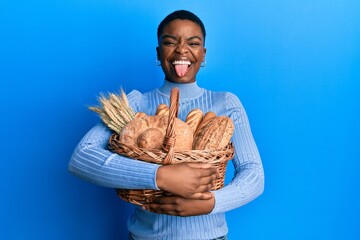 Young african american woman holding wicker basket with bread sticking tongue out happy with funny expression.