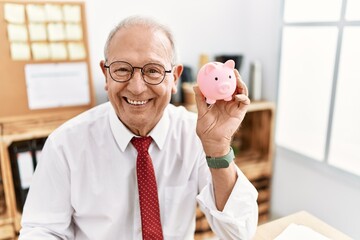 Senior business man holding piggy bank looking positive and happy standing and smiling with a confident smile showing teeth