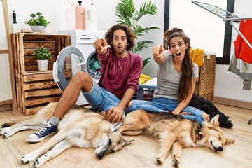 Sticker - Young hispanic couple doing laundry with dogs pointing with finger surprised ahead, open mouth amazed expression, something on the front